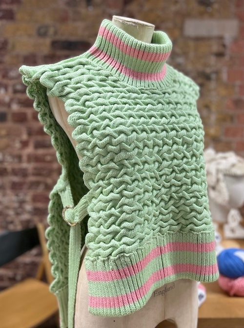Green knitted vest from the REVIVAL capsule collection by ACIEN x WOVEN BEYOND, crafted from French Merino D’Arles wool and Himalayan nettle. Features ACIEN's signature freckle knit texture, a turtle neck and chunky rib with two pink stripes, and open sides with self ties.