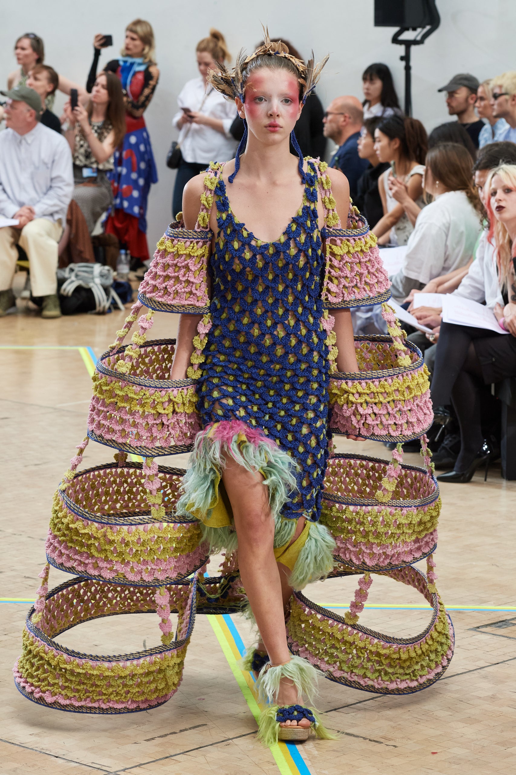 Look 2, PERETA from REGENERATIVE FOLKLORE, showcased at the Central Saint Martins press catwalk. Green and blue crochet dress made from pineapple yarn with green fur detail at the bottom. Paired with a green and pink crochet structure made from nettle yarn and dry branches from Southern Spain, a green crochet headpiece and green fur implanted open-toe wedges with blue crochet carnation details.