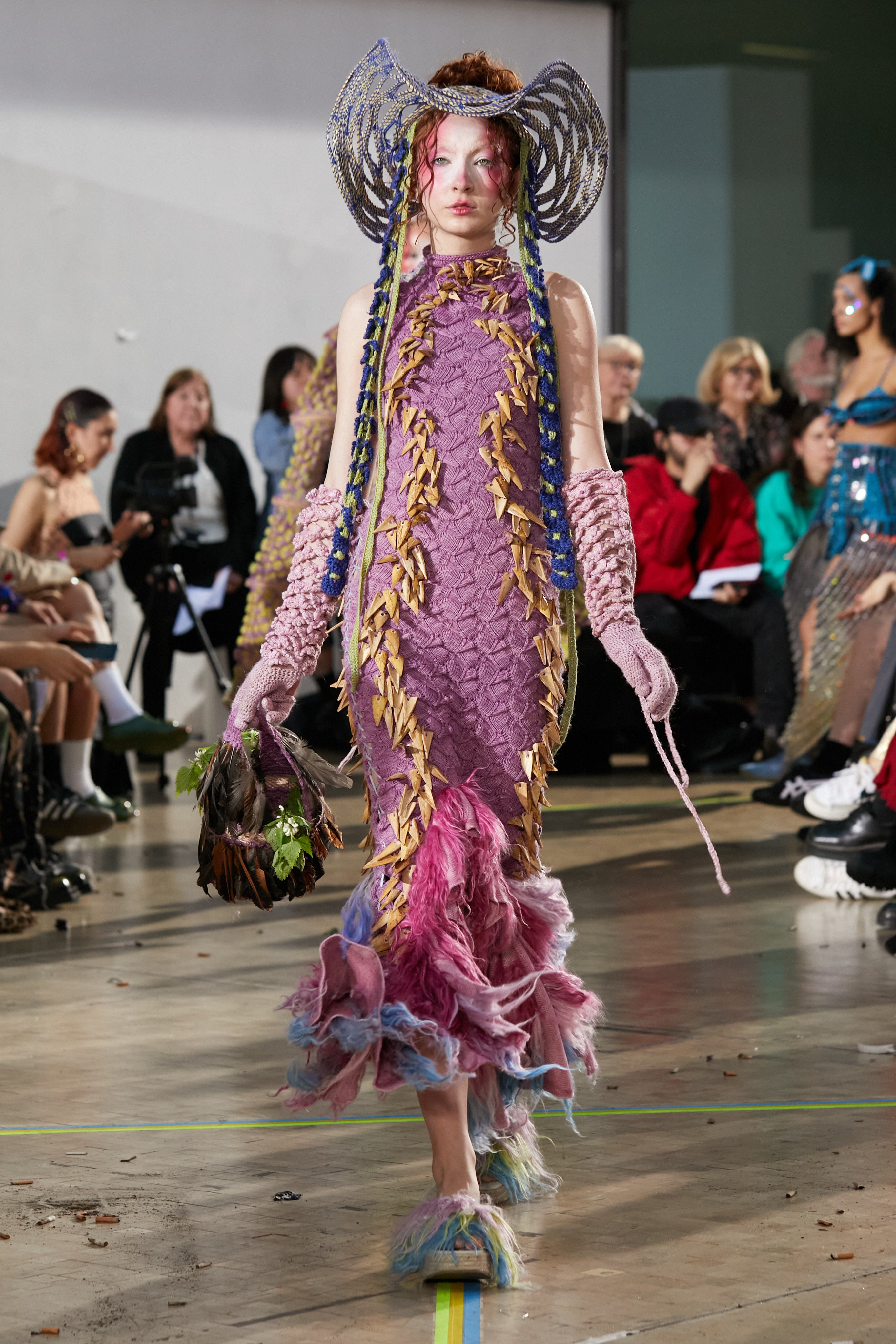 Look 3, TABERNAS from REGENERATIVE FOLKLORE, showcased at the Central Saint Martins press catwalk. Pink knit dress made from nettle yarn, dyed from invasive plants adorned with hand cut Almería canes. Paired with the TABERNAS hat, meticulously woven using ancestral techniques with dry branches sourced from the mountains of Southern Spain. Adorned with blue and green crocheted pineapple and tencel flower straps. Also paired with pink crochet gloves and pink, blue and green fur implanted open-toe wedges.