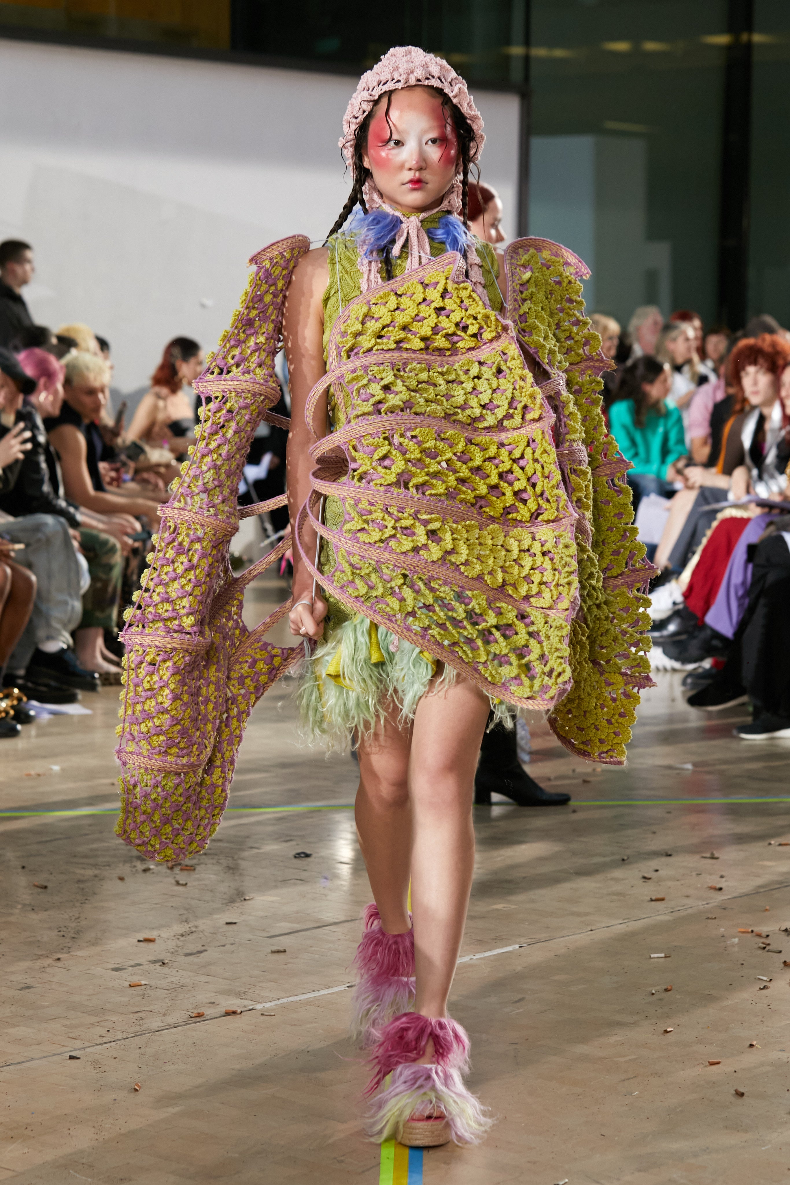 Look 4, ABANICOS from REGENERATIVE FOLKLORE, showcased at the Central Saint Martins press catwalk. Green and pink modular crochet structure made from nettle yarn, dyed with invasive plants and woven dry branches from Southern Spain over a short green knitted dress. Paired with pink crochet hat dyed with bacteria and dark and light pink fur-implanted open-toe wedges.