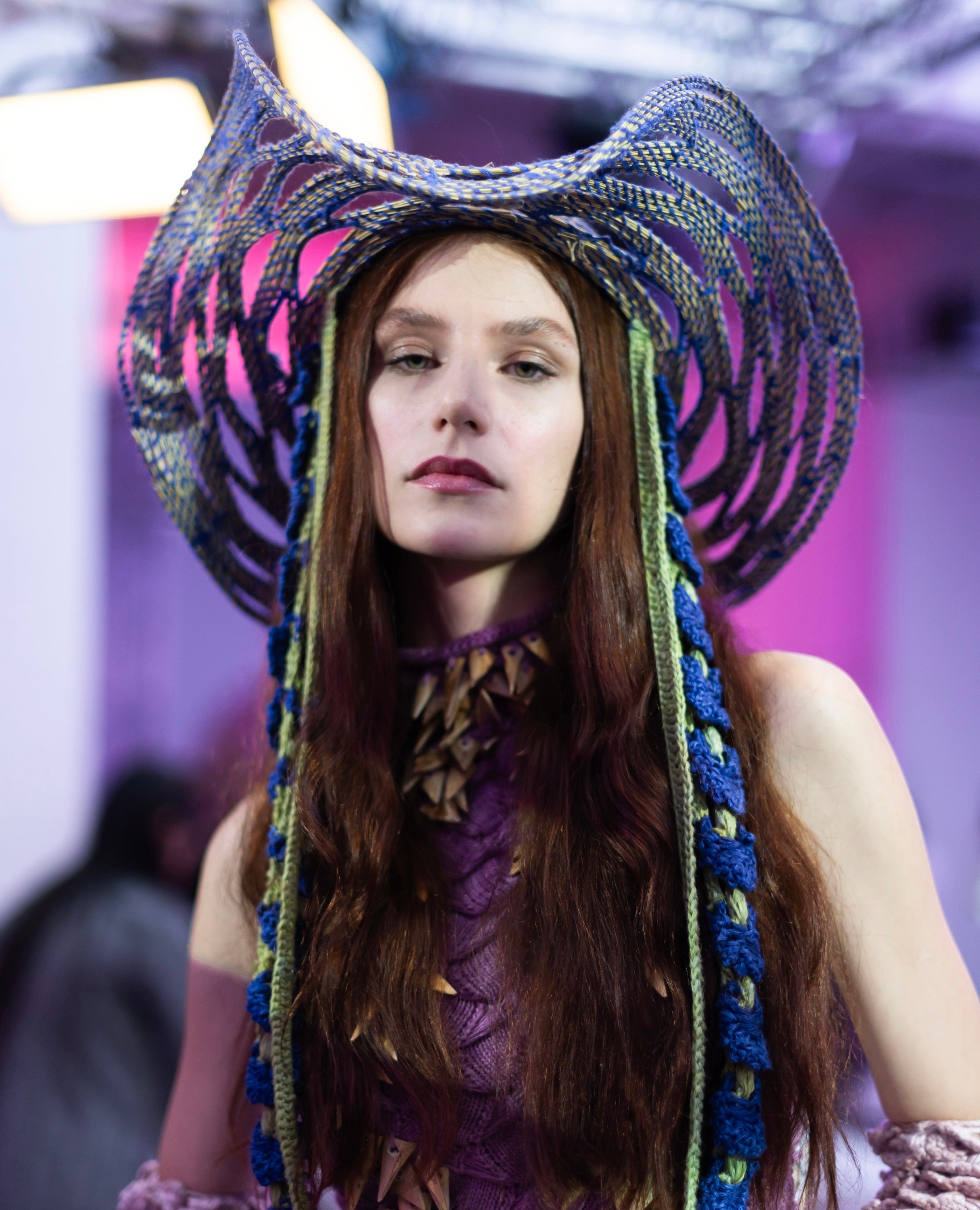 Close-up of Tina Kolodiy wearing Look 3, TABERNAS, from REGENERATIVE FOLKLORE at London Fashion Week 2023. Pink knit dress made from nettle yarn, dyed from invasive plants adorned with hand cut Almería canes. Paired with the TABERNAS hat, meticulously woven using ancestral techniques with dry branches sourced from the mountains of Southern Spain. Adorned with blue and green crocheted pineapple and tencel flower straps.