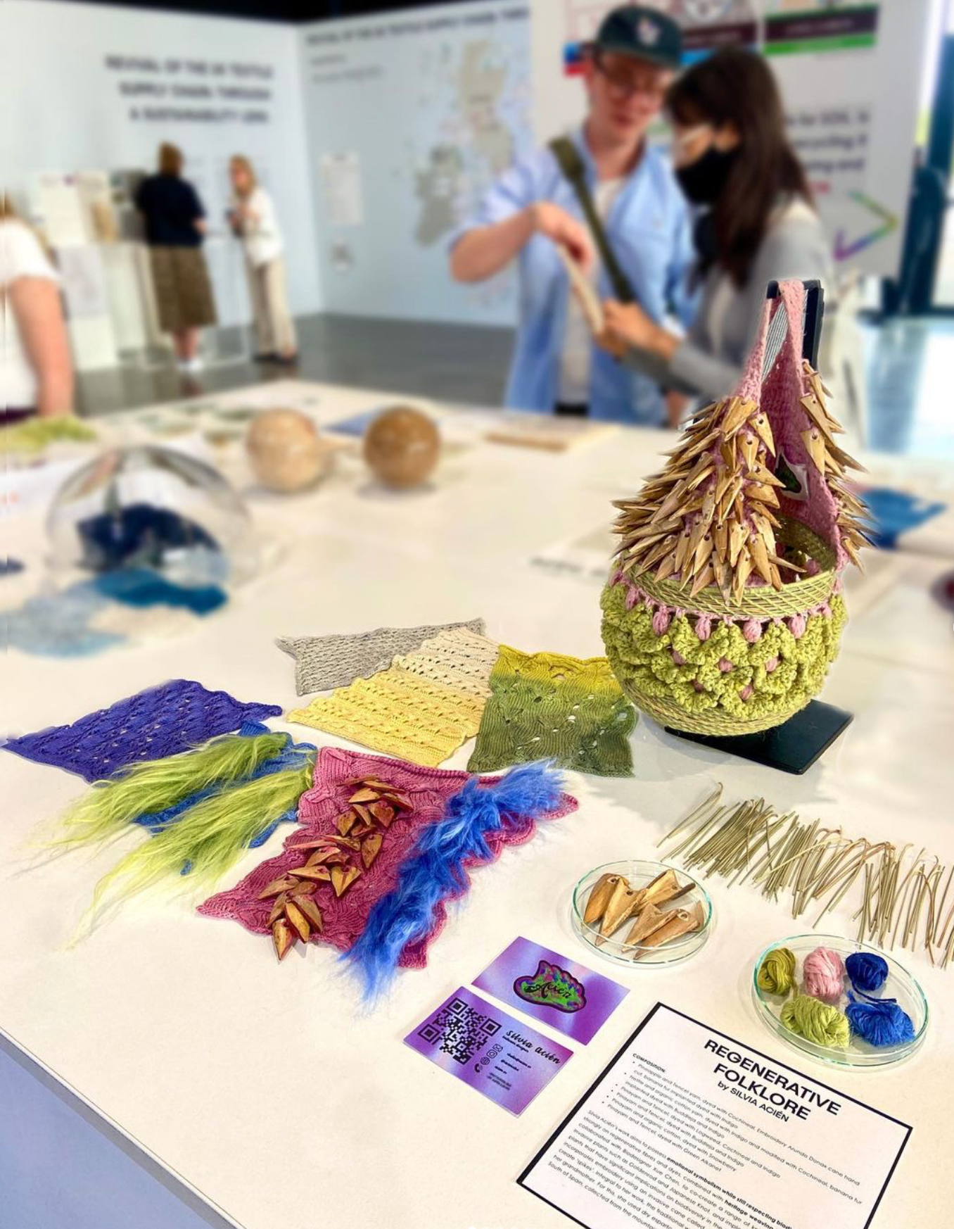 REGENERATIVE FOLKLORE at The Future Fabric Expo. Table showcasing REGENERATIVE FOLKLORE knitted textile samples,green and pink crochet bag adored with hand cut Almería canes, dry branches from Southern Spain samples and two petri dishes containing hand cut Almería canes and naturally dyed pineapple and nettle yarn samples.